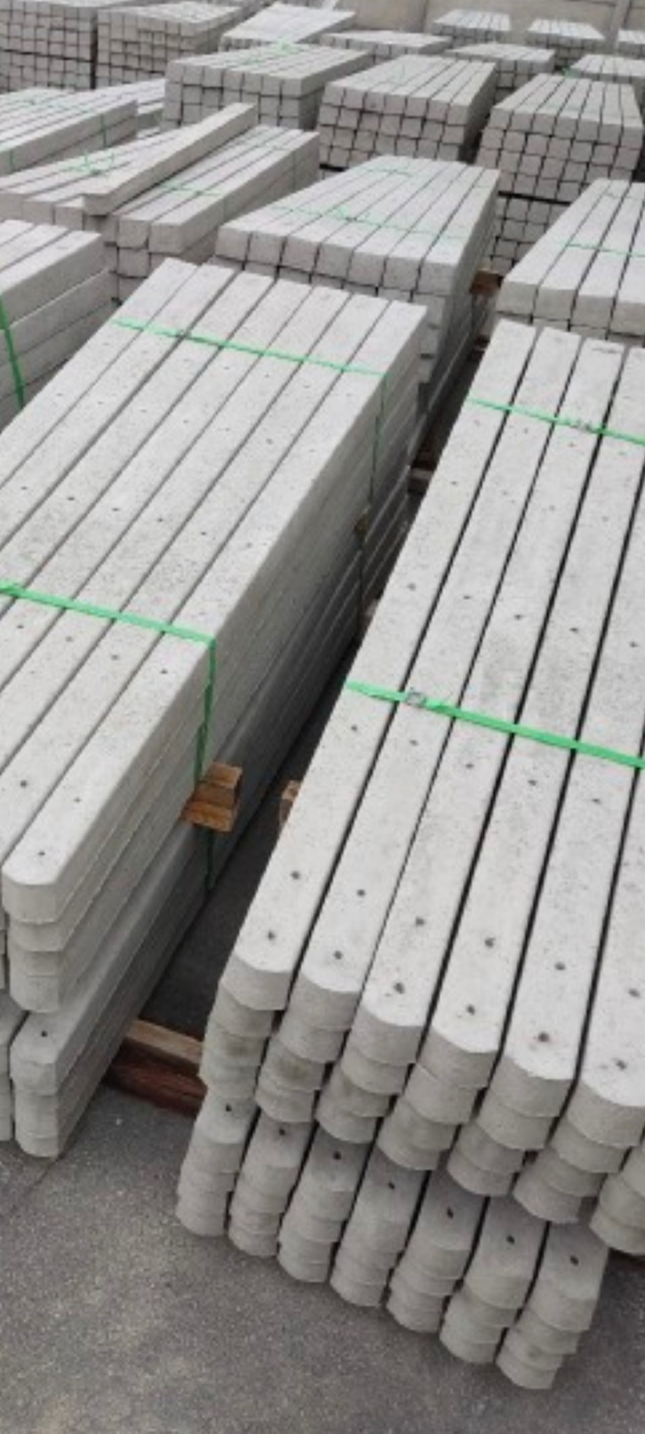 Szare betonowe słupki do siatki ogrodzeniowej ułożone w stosy, spięte zielonymi taśmami, widoczne otwory montażowe.