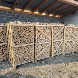 Rząd drewnianych skrzyń wypełnionych pociętym drewnem kominkowym, ułożonych pod zadaszeniem z szarej cegły.