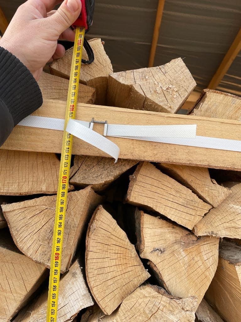 Pomiar metrem zwiniętym ułożonego stosu pociętego drewna opałowego, spiętego białą taśmą, widok z bliska.