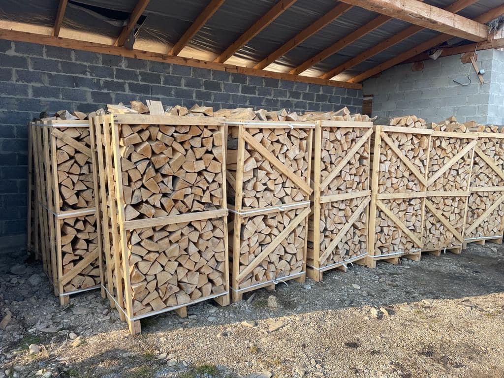 Rząd drewnianych skrzyń wypełnionych pociętym drewnem kominkowym, ułożonych pod zadaszeniem z szarej cegły.