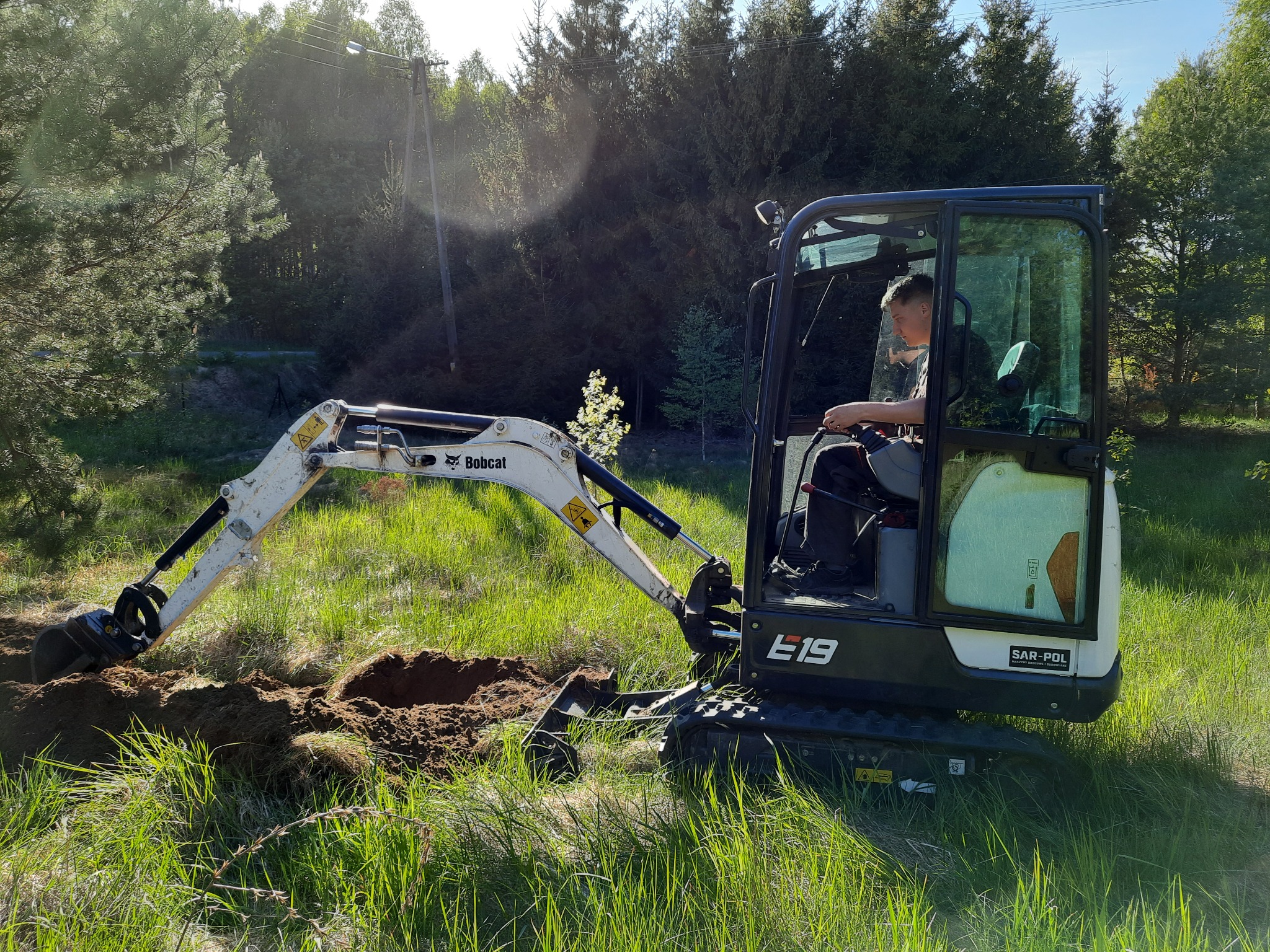 Operator w minikoparce Bobcat E19 wykonuje wykop w zarośniętym terenie, widoczne świeżo wykopane zagłębienie w ziemi i otaczająca roślinność.