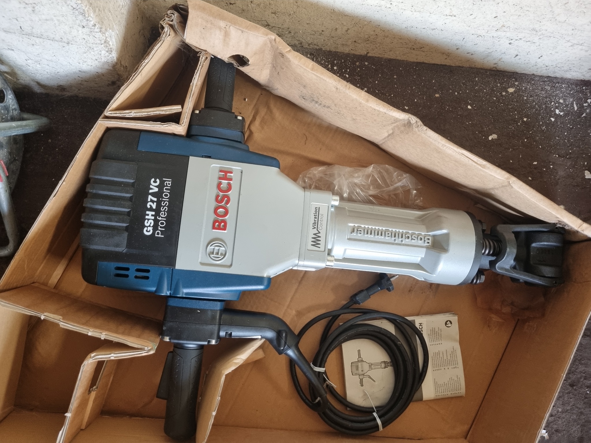 Młot udarowy Bosch GSH 27 VC Professional w kartonowym opakowaniu, widoczny kabel zasilający i instrukcja obsługi.