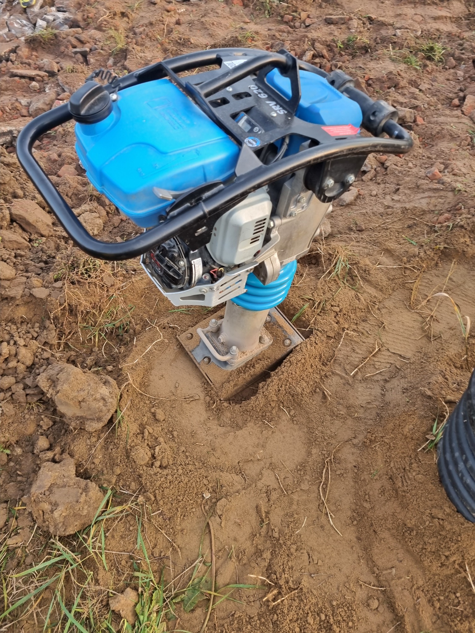 Zagęszczarka stopowa z niebieskim zbiornikiem paliwa zagęszcza glebę w wykopie, widoczne ślady pracy i grudki ziemi.