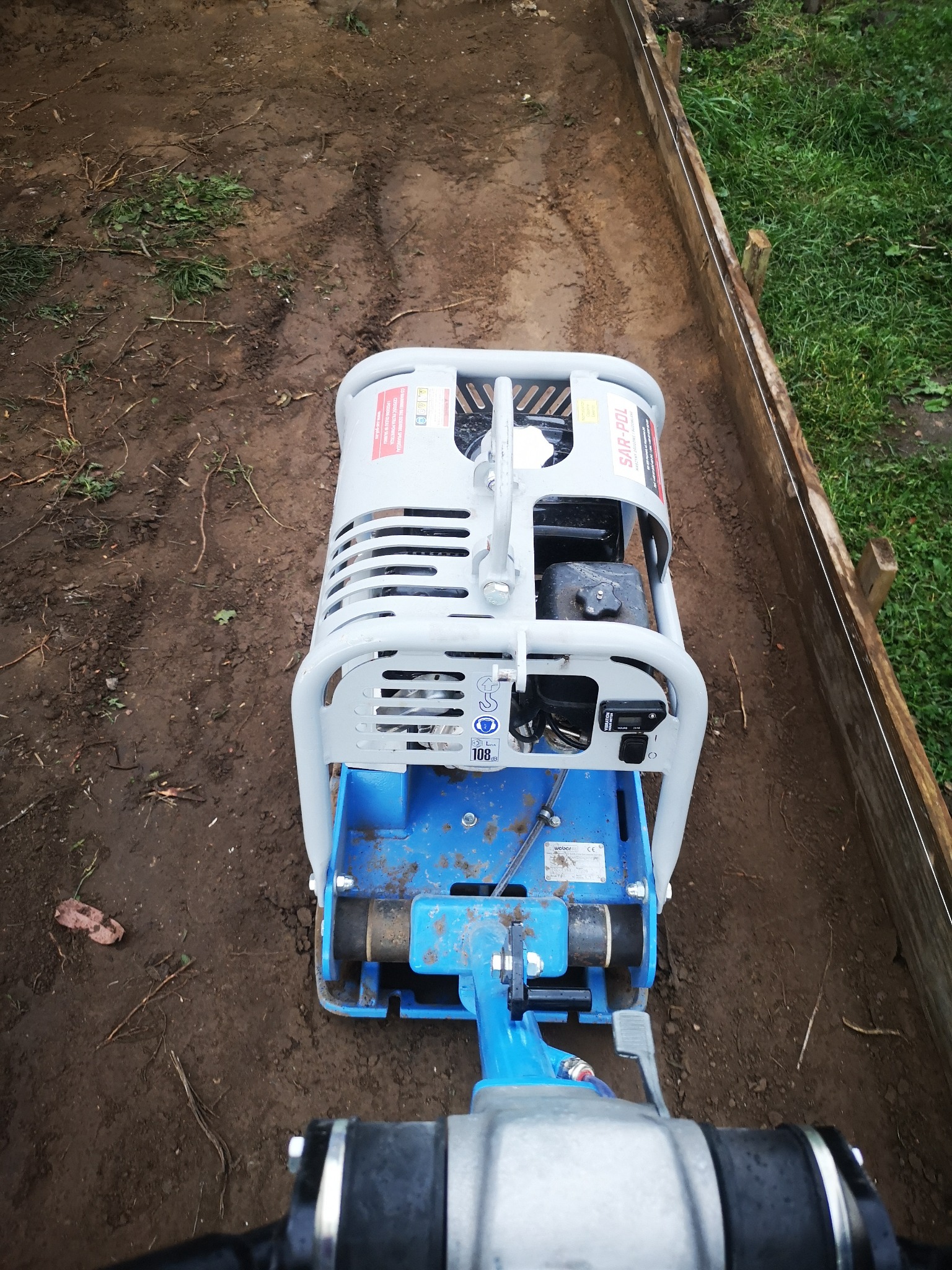 Zagęszczarka płytowa, niebiesko-szara, ubija ziemię w wyznaczonym drewnianym korycie, widoczne ślady po przejazdach, świeżo ubita ziemia.