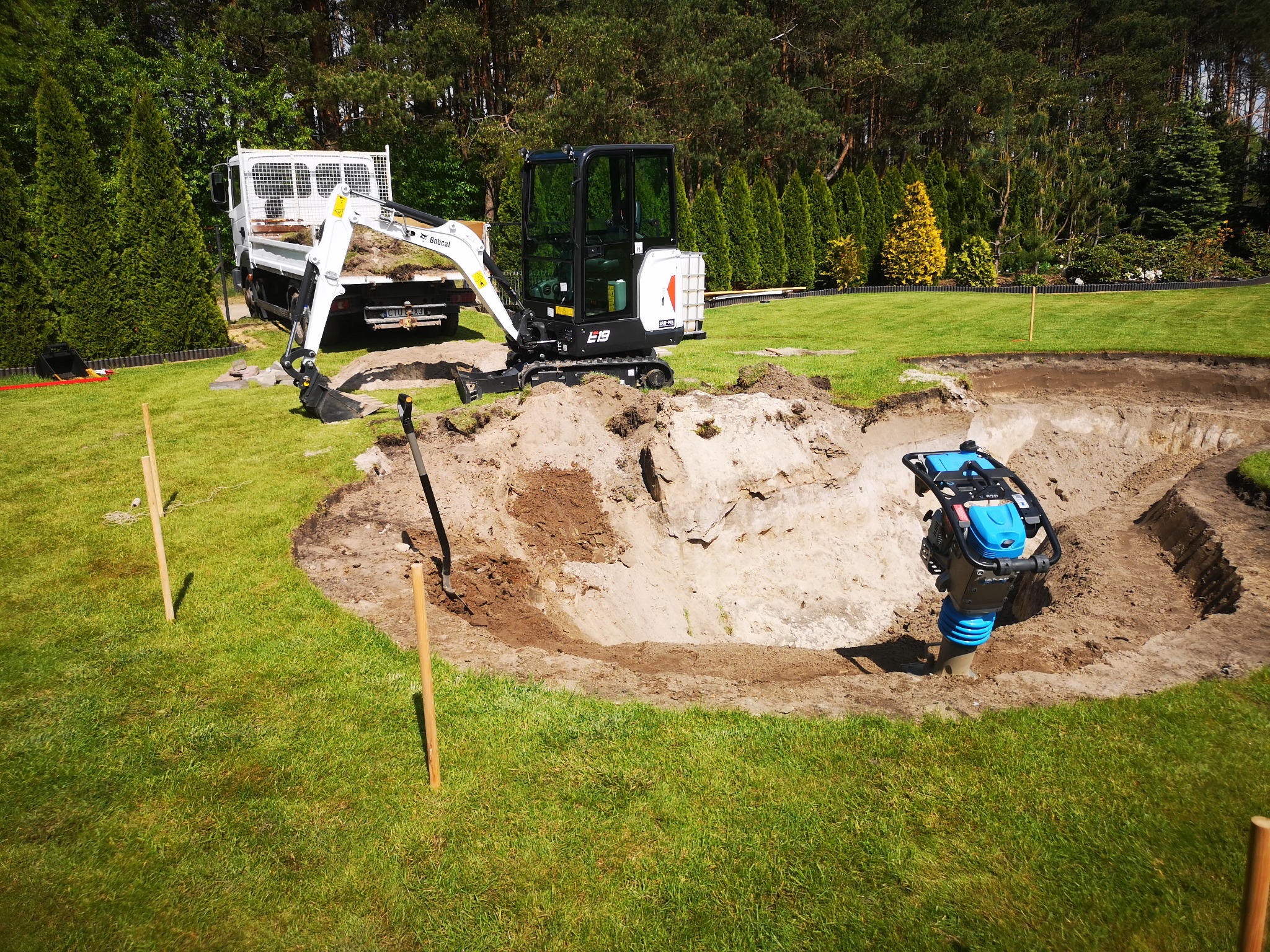 Koparka Bobcat E19 i ubijak wibracyjny w wykopie o nieregularnym kształcie na trawniku, obok zaparkowany samochód dostawczy, w tle drzewa iglaste i liściaste.