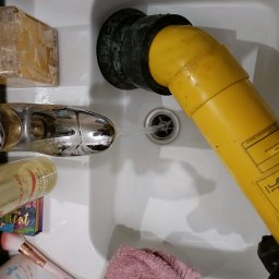 Dom Serwis - Żółta pompa z czarną gumową uszczelką w zlewie, strumień wody z kranu skierowany do otworu w pompie, obok kosmetyki i ręcznik.