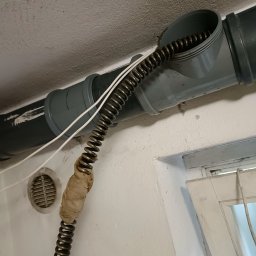 Dom Serwis - Sprężyna do czyszczenia rur wprowadzone do szarej rury kanalizacyjnej, owinięta taśmą w połowie długości, widoczny fragment wentylacji i okna w tle.