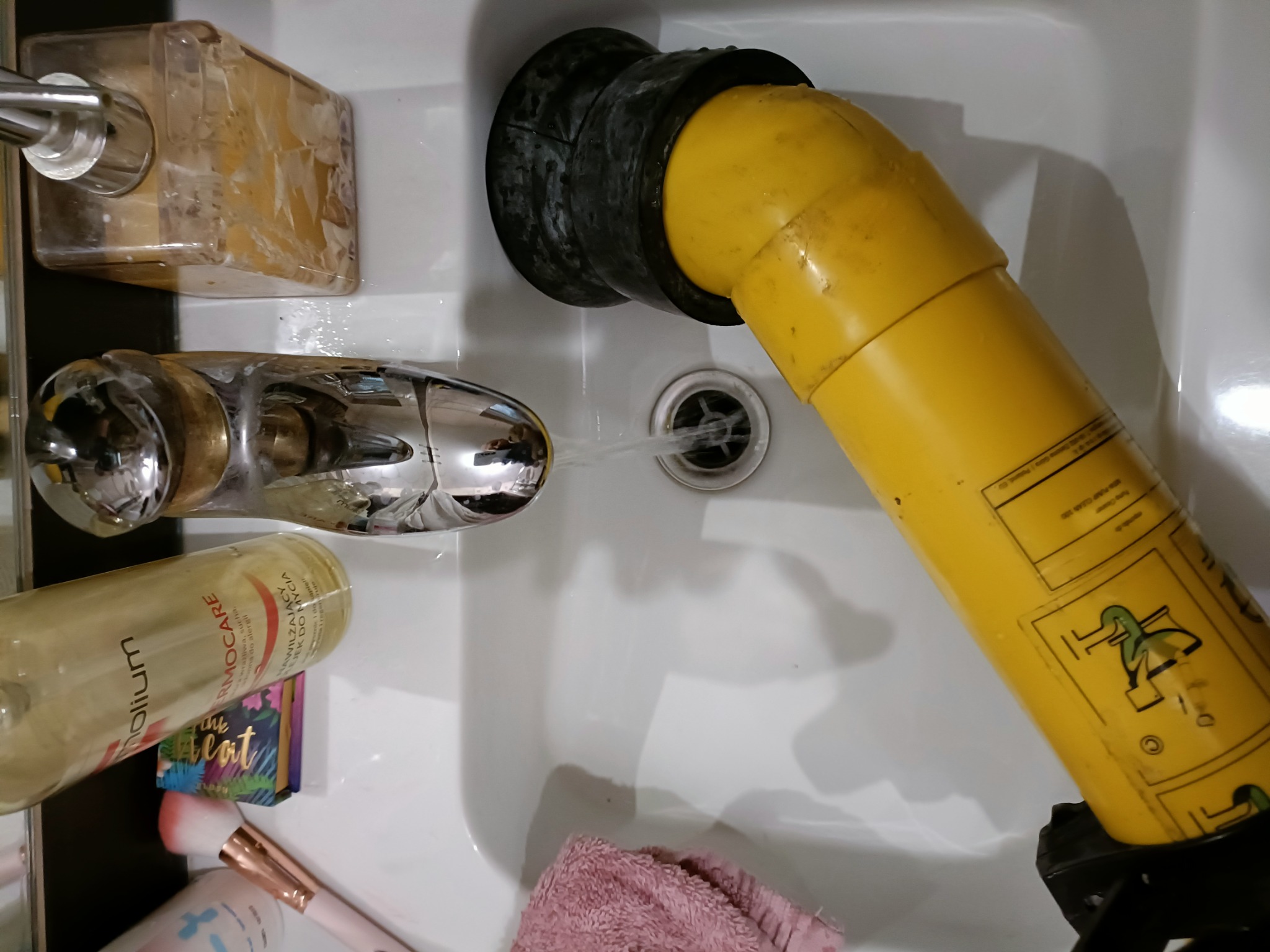 Żółta pompa z czarną gumową uszczelką w zlewie, strumień wody z kranu skierowany do otworu w pompie, obok kosmetyki i ręcznik.