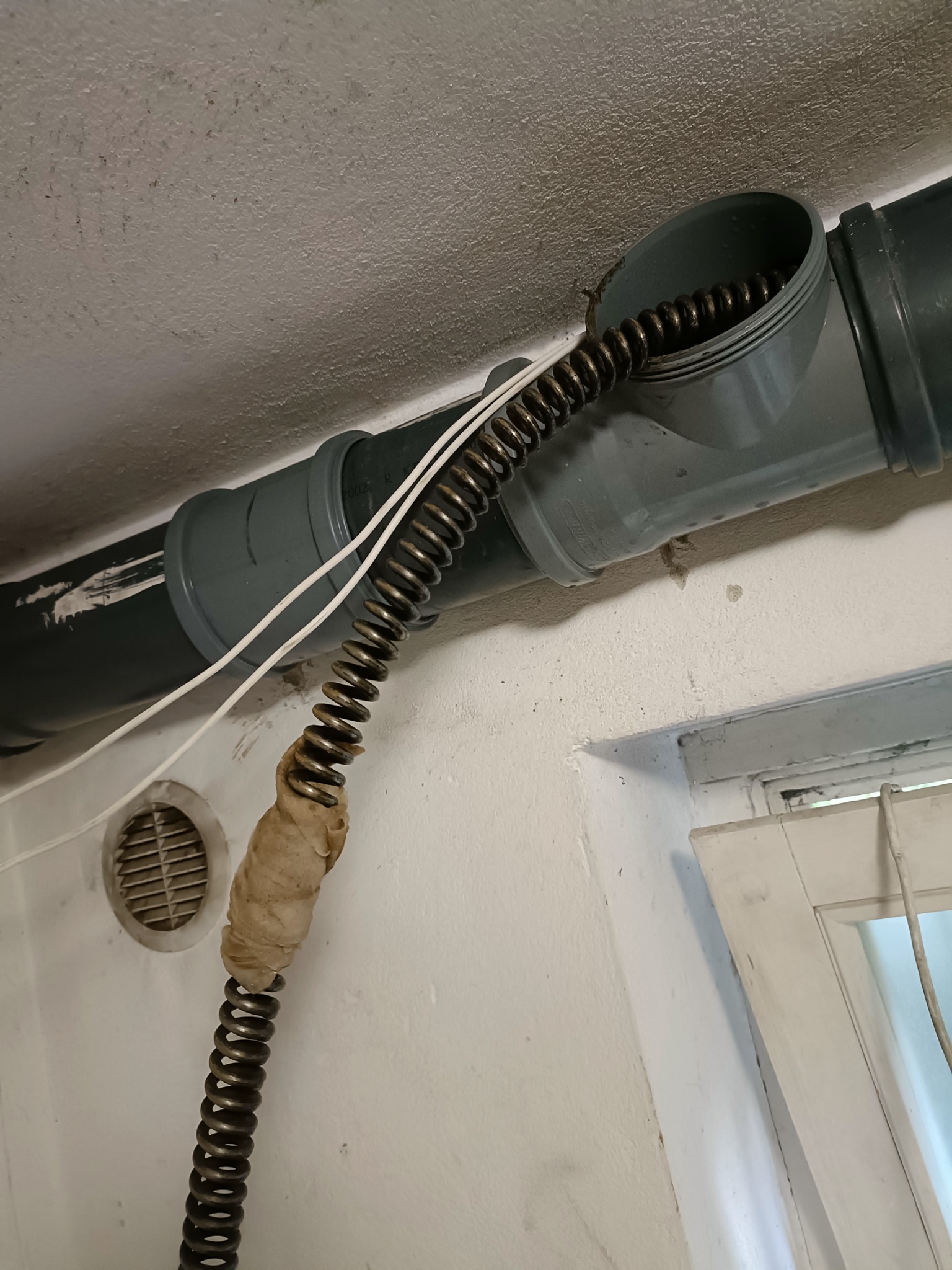 Sprężyna do czyszczenia rur wprowadzone do szarej rury kanalizacyjnej, owinięta taśmą w połowie długości, widoczny fragment wentylacji i okna w tle.