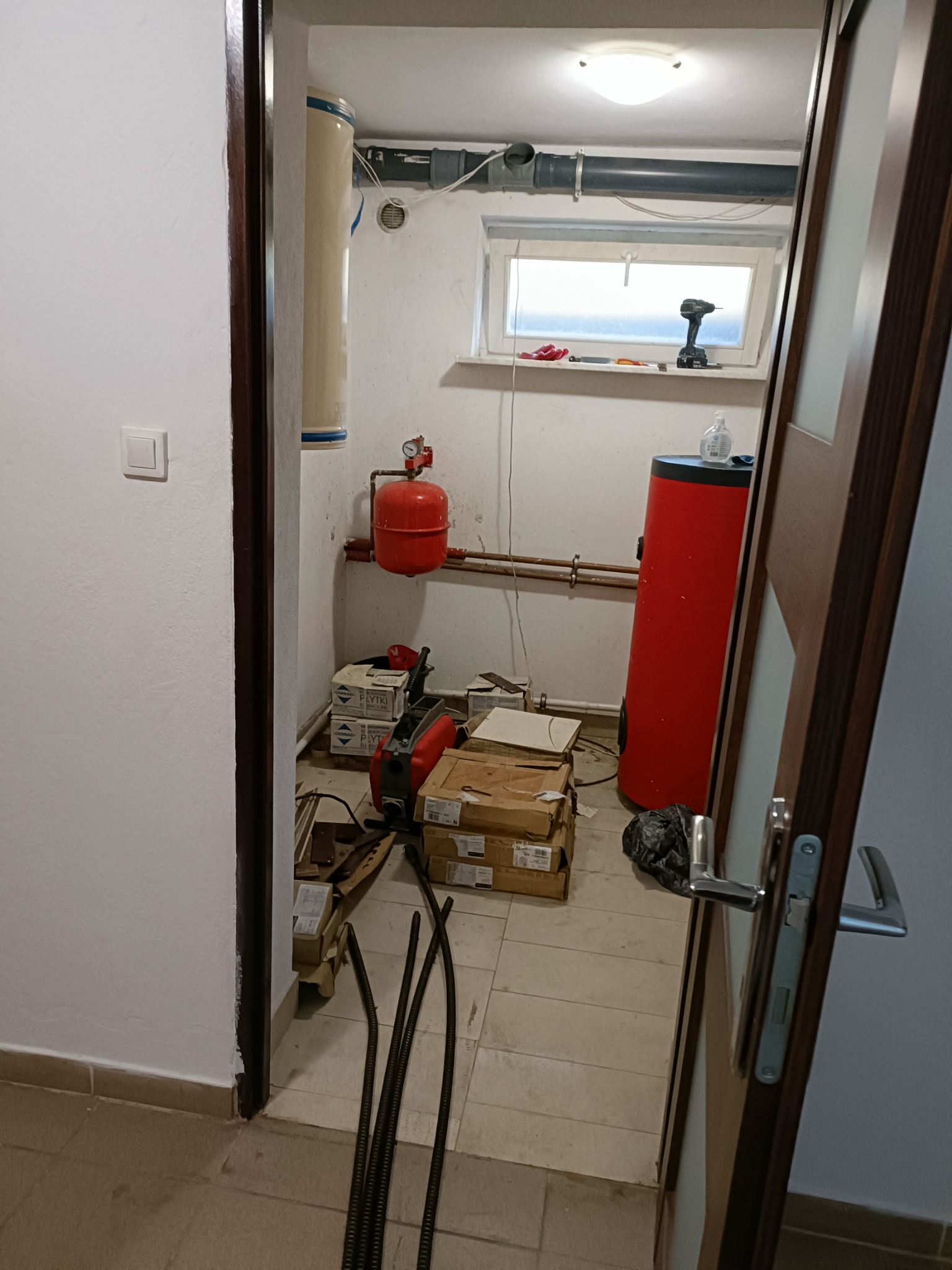 Piwnica z dwoma zbiornikami na wodę (czerwony i beżowy), skrzynki z płytkami, narzędzia i przewody leżące na podłodze, widok zza otwartych drzwi.