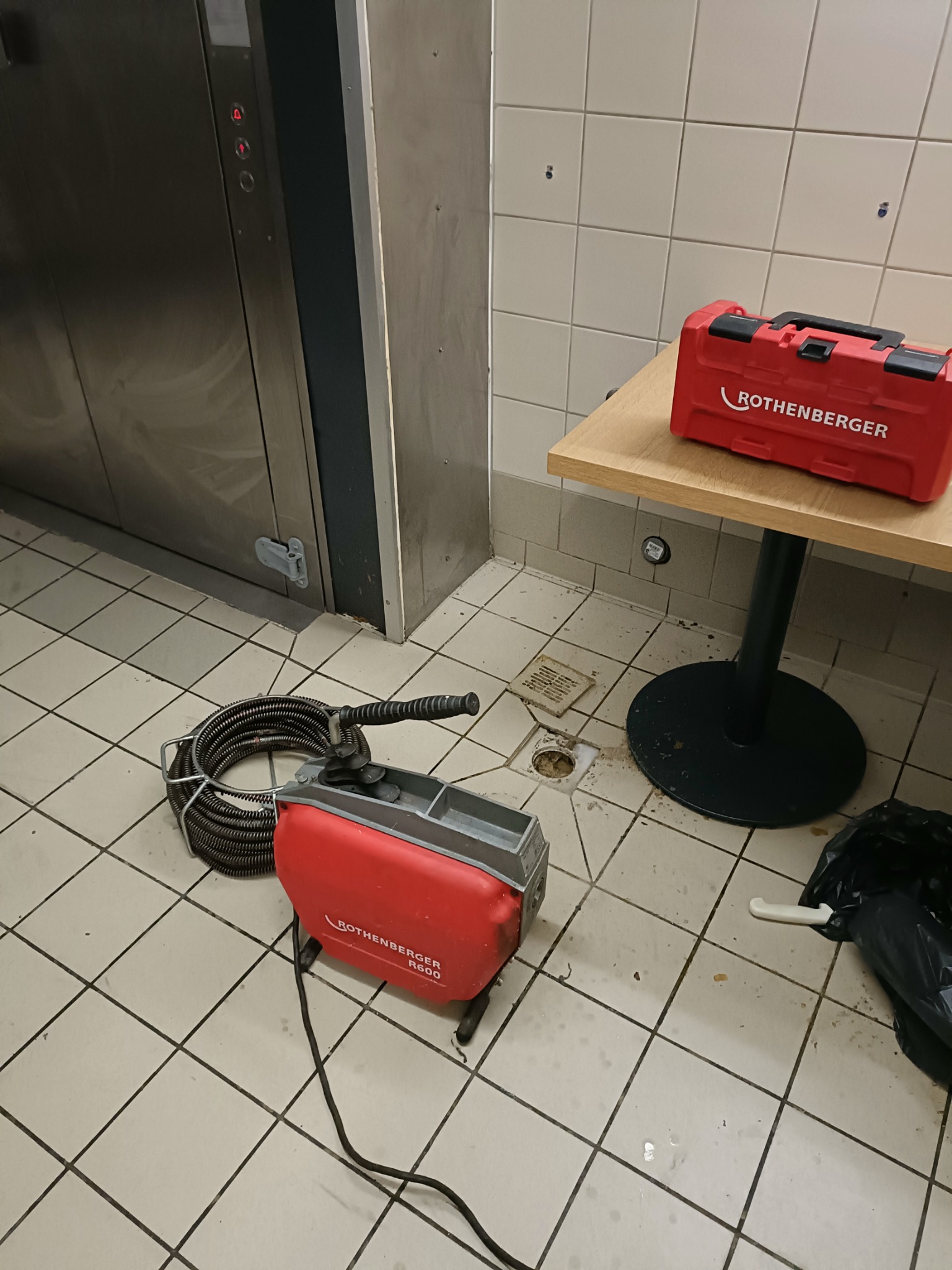 Elektryczna sprężyna do udrażniania rur Rothenberger R600 obok otwartego odpływu podłogowego w pobliżu windy i czerwonej skrzynki narzędziowej Rothenberger na stoliku.