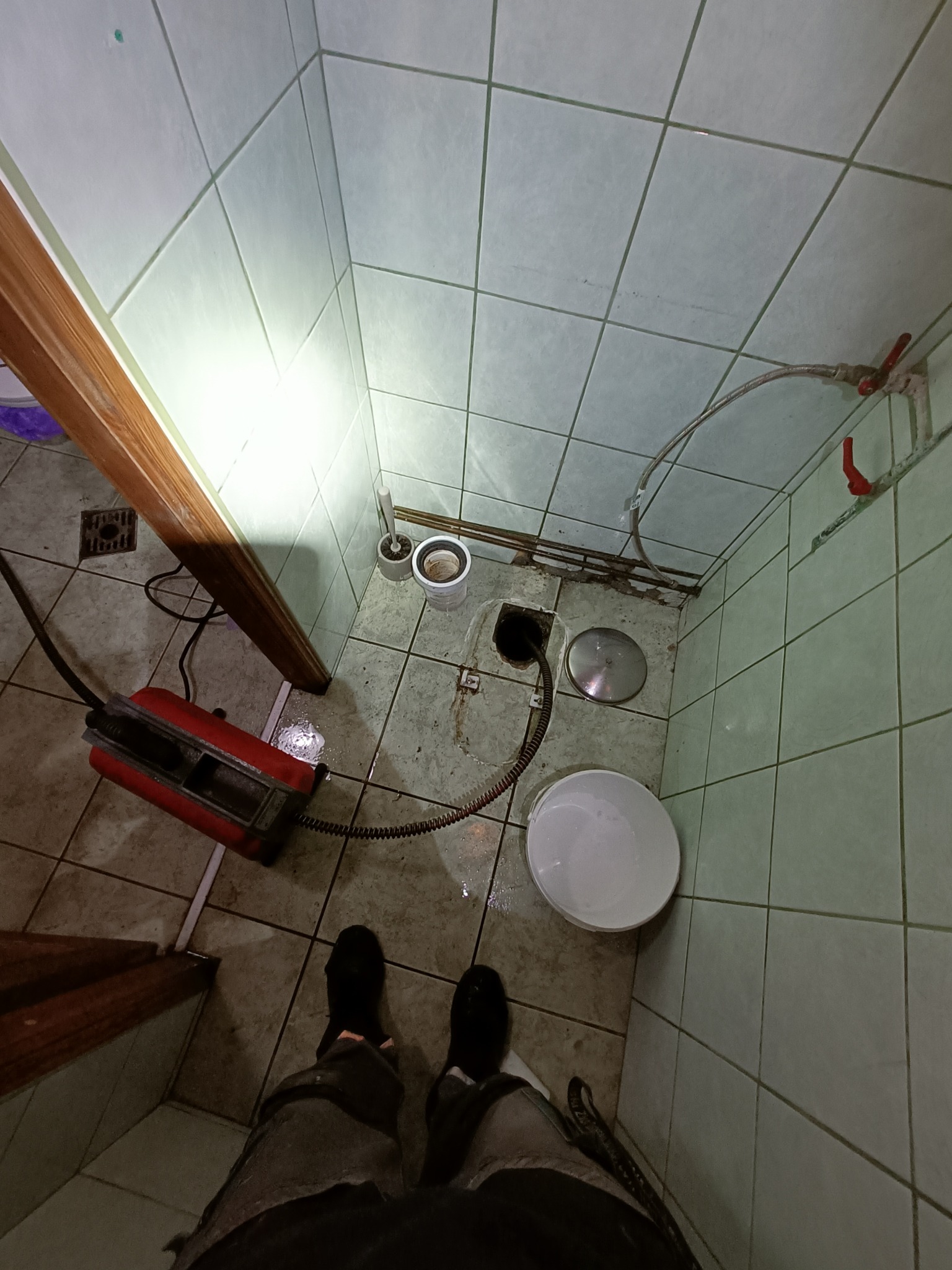 Widok z góry na łazienkę z kwadratowymi płytkami, gdzie widać czerwone urządzenie do czyszczenia rur, białe wiadro i otwarty odpływ podłogowy z ciemną zawartością w trakcie pracy.