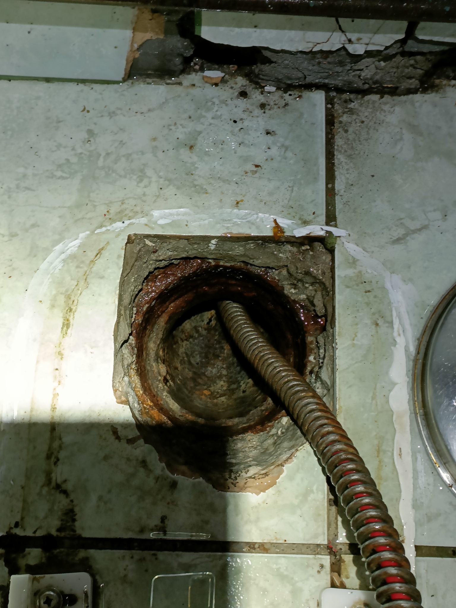 Zardzewiała rura kanalizacyjna z wprowadzoną sprężyną do udrażniania, widoczne uszkodzenia płytek i ślady wilgoci.