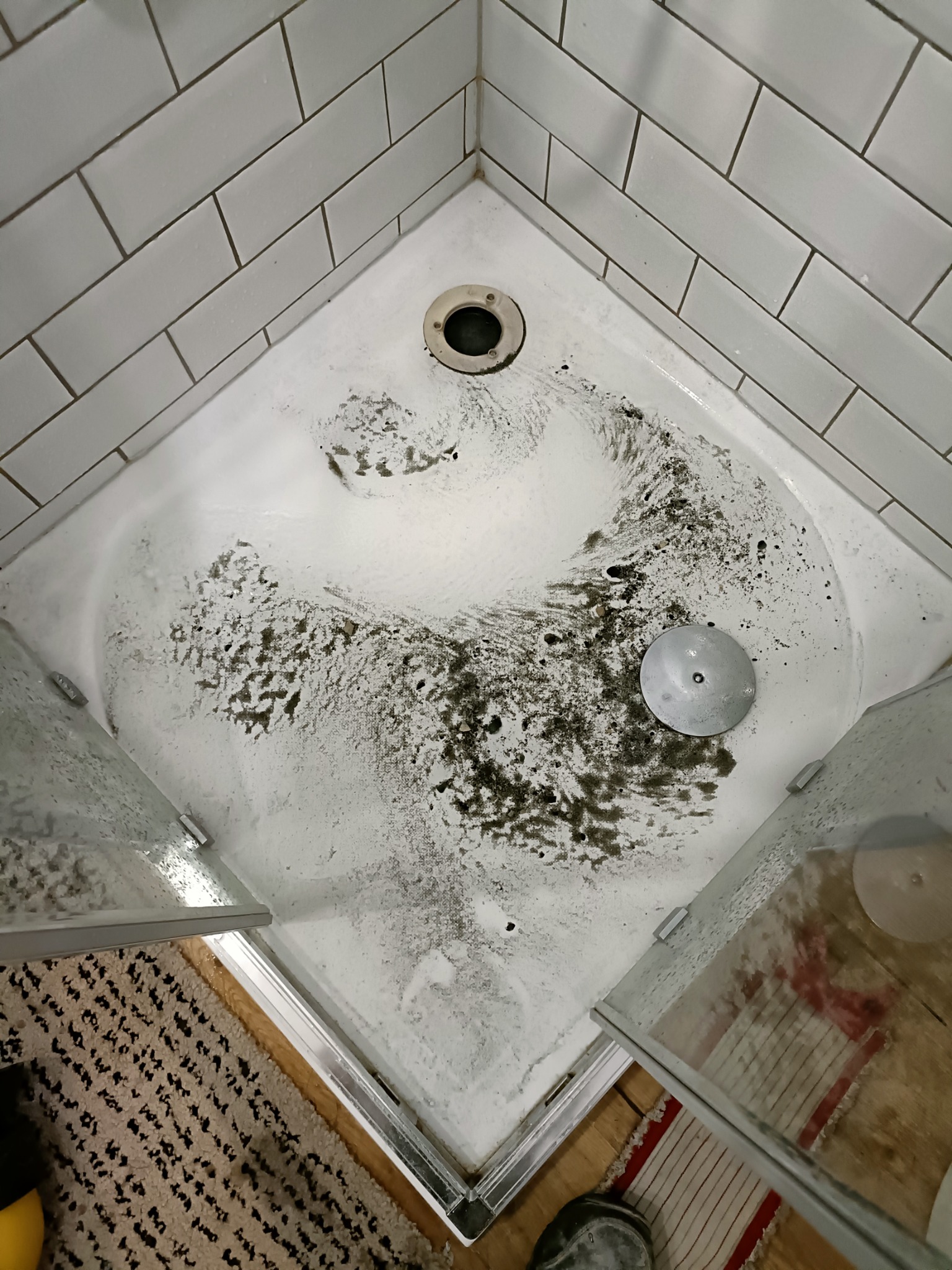 Brudny brodzik po czyszczeniu, widoczne resztki zanieczyszczeń i piany, białe kafelki na ścianach, metalowy odpływ w rogu i częściowo otwarte szklane drzwi kabiny.