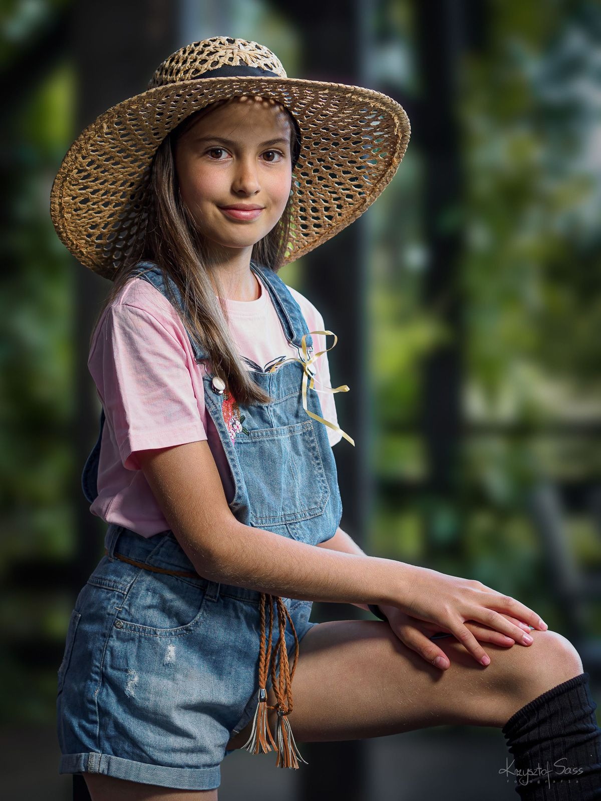 Portret uśmiechniętej nastolatki w słomkowym kapeluszu, różowej koszulce i dżinsowych ogrodniczkach na tle zieleni, stylizowane ujęcie.
