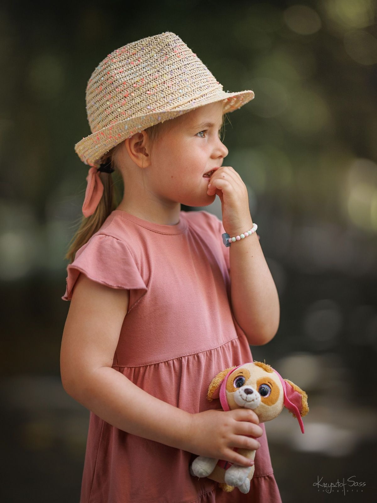 Portret dziewczynki w słomkowym kapeluszu i różowej sukience, trzymającej pluszowego pieska i spoglądającej w dal, z delikatnym rozmyciem tła.