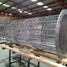 Szkielet wymiennika ciepła z rur i okrągłych płyt perforowanych, wsparty na drewnianych belkach w hali produkcyjnej.