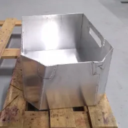 Prostopadłościenny pojemnik ze spawanych blach, z uchwytami po bokach, umieszczony na drewnianej palecie w warsztacie.