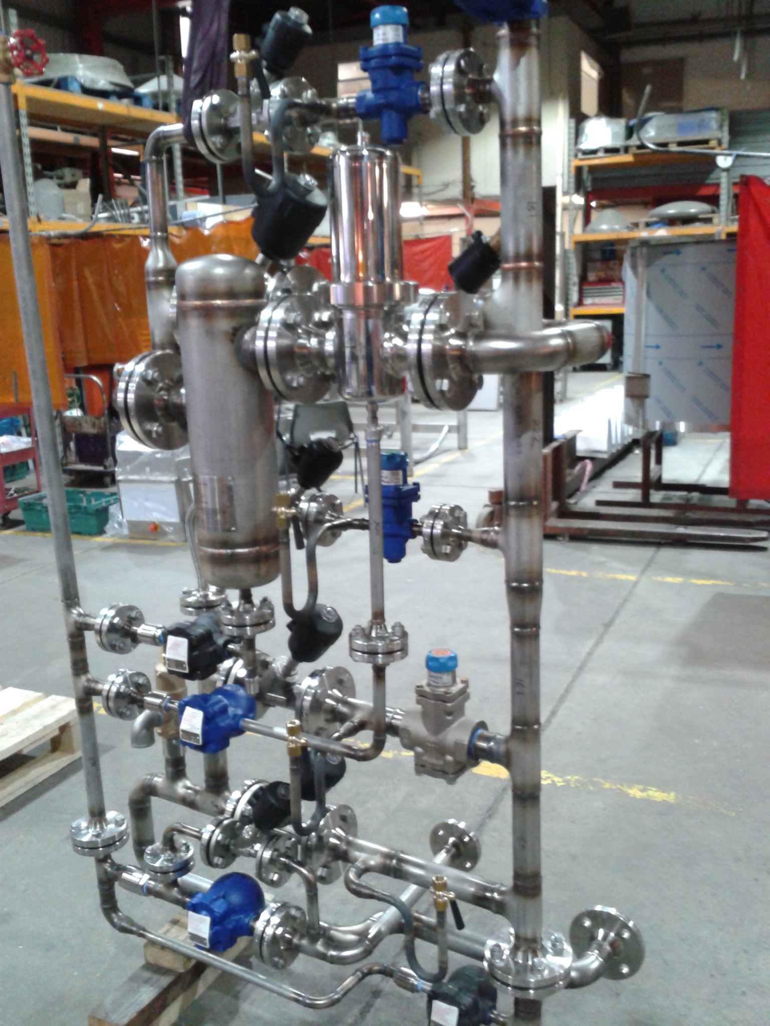Złożona instalacja przemysłowa z rur i zaworów ze stali nierdzewnej, widoczne liczne łączenia kołnierzowe i elementy automatyki, ustawiona na drewnianych paletach w hali produkcyjnej.