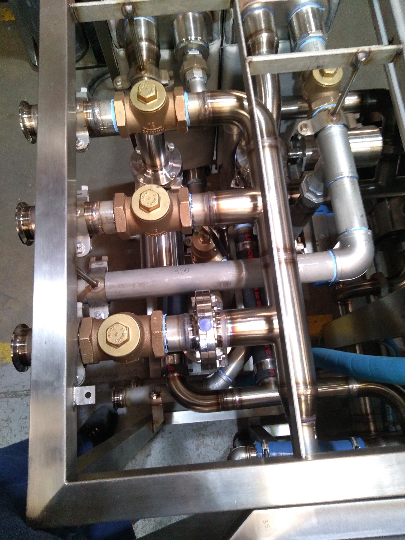 Kompleksowy system rur i zaworów ze stali nierdzewnej i mosiądzu, z widocznymi spawami i niebieskimi elementami, zamontowany w metalowej ramie.