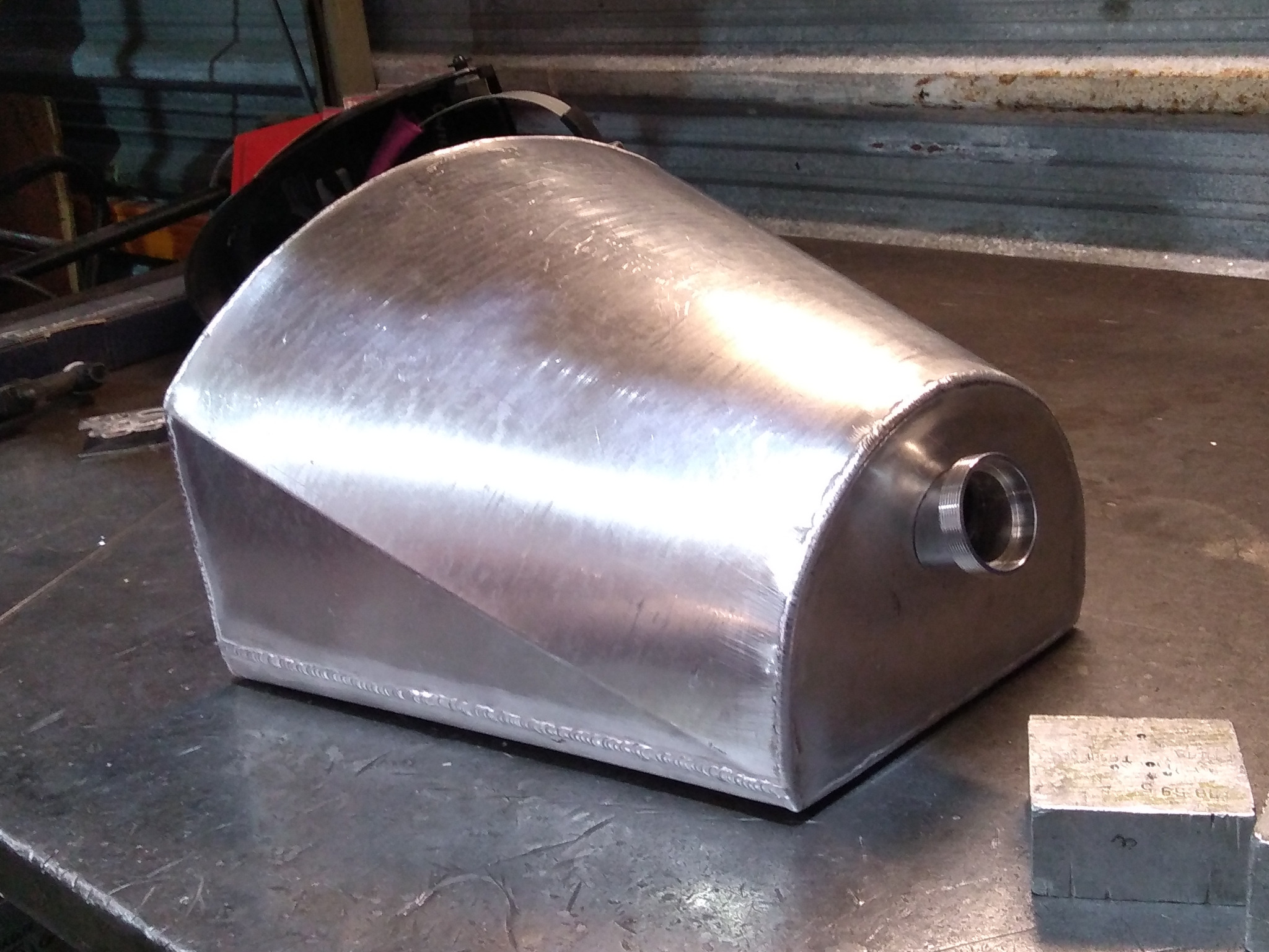 Metalowy zbiornik o nietypowym kształcie, spawany, z widocznym otworem z gwintem, leżący na metalowym stole w warsztacie, obok leży metalowy prostopadłościan.