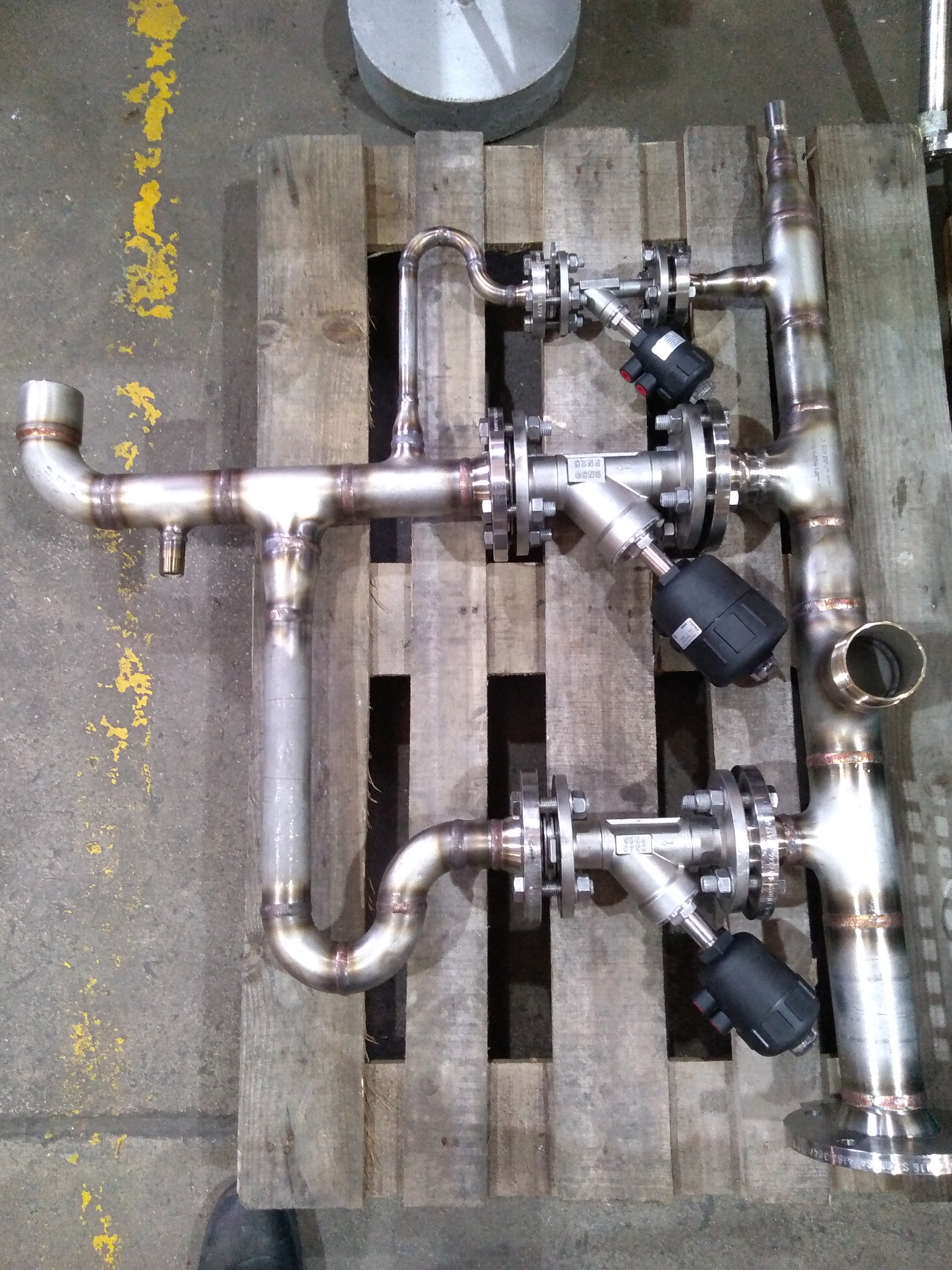 Złożony system rur ze stali nierdzewnej z zaworami i przyłączami kołnierzowymi, leżący na drewnianej palecie w warsztacie.