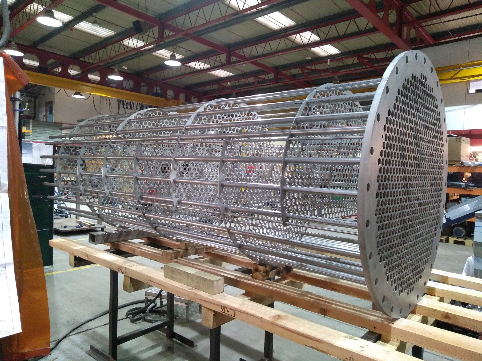 Szkielet wymiennika ciepła z rur i okrągłych płyt perforowanych, wsparty na drewnianych belkach w hali produkcyjnej.