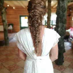 Upięcie ślubne: brązowe włosy spięte w skomplikowany kok z luźno opadającymi lokami na plecach, kobieta w białej koronkowej sukni z odkrytymi plecami, widok z tyłu, rustykalne wnętrze z drewnianymi...