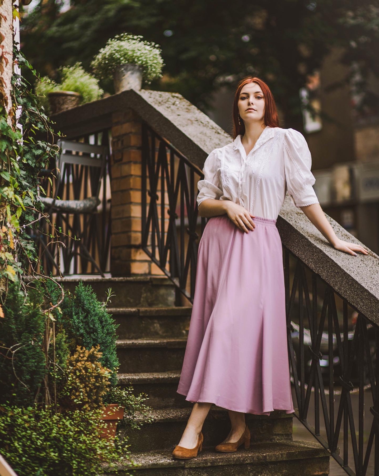 Kobieta z rudymi włosami w białej bluzce i długiej różowej spódnicy pozuje na kamiennych schodach z metalową balustradą, otoczonych zielenią.