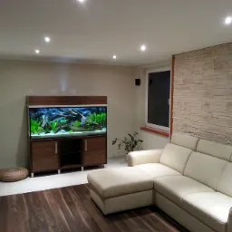 Salon z dekoracyjnym akwarium w zabudowie z ciemnego drewna, podświetlone punktowo, z roślinami i korzeniami wewnątrz, obok beżowa sofa i ściana wyłożona kamieniem dekoracyjnym.