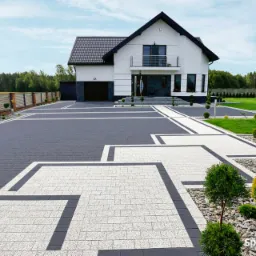 Nowoczesny podjazd do domu z kostki brukowej w odcieniach szarości i bieli, z dekoracyjnymi obwódkami i nasadzeniami z kulistych krzewów.