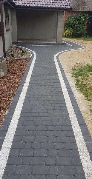 Ułożona, ciemnoszara kostka brukowa z białymi obrzeżami tworząca ścieżkę rozwidlającą się w kierunku wejścia do budynku i trawnika.