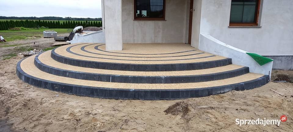 Półokrągłe schody wejściowe z kostki brukowej w kolorze piaskowym i grafitowym, prowadzące do domu z surowym wykończeniem elewacji, widoczne ślady prac ziemnych.