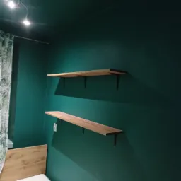 Dwie drewniane półki zamontowane na ścianie pomalowanej na ciemnozielony kolor, widoczny fragment łóżka z drewnianym zagłówkiem i zasłona w liście.