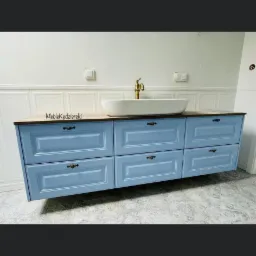 Niebieska szafka łazienkowa pod umywalkę nablatową z drewnianym blatem i mosiężną baterią, wisząca na ścianie z białą boazerią.