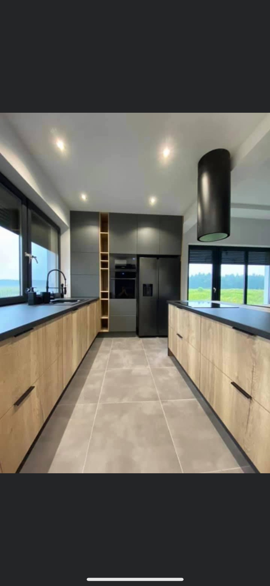 Nowoczesna kuchnia w stylu minimalistycznym z drewnianymi frontami szafek i czarnymi blatami, oświetlona punktowymi lampami sufitowymi i cylindrycznym okapem nad wyspą kuchenną, widok na zewnątrz...