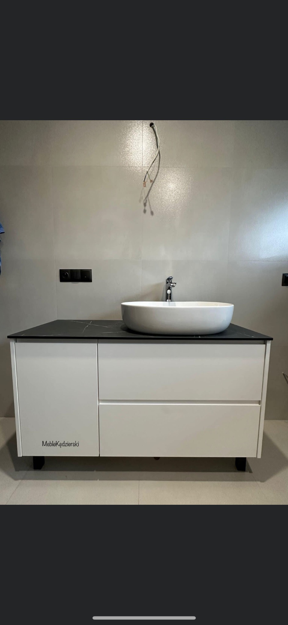 Biała szafka łazienkowa pod umywalkę z czarnym blatem, widoczne logo 'MebleKędzierski' na froncie, niedokończone podłączenie elektryczne na ścianie.