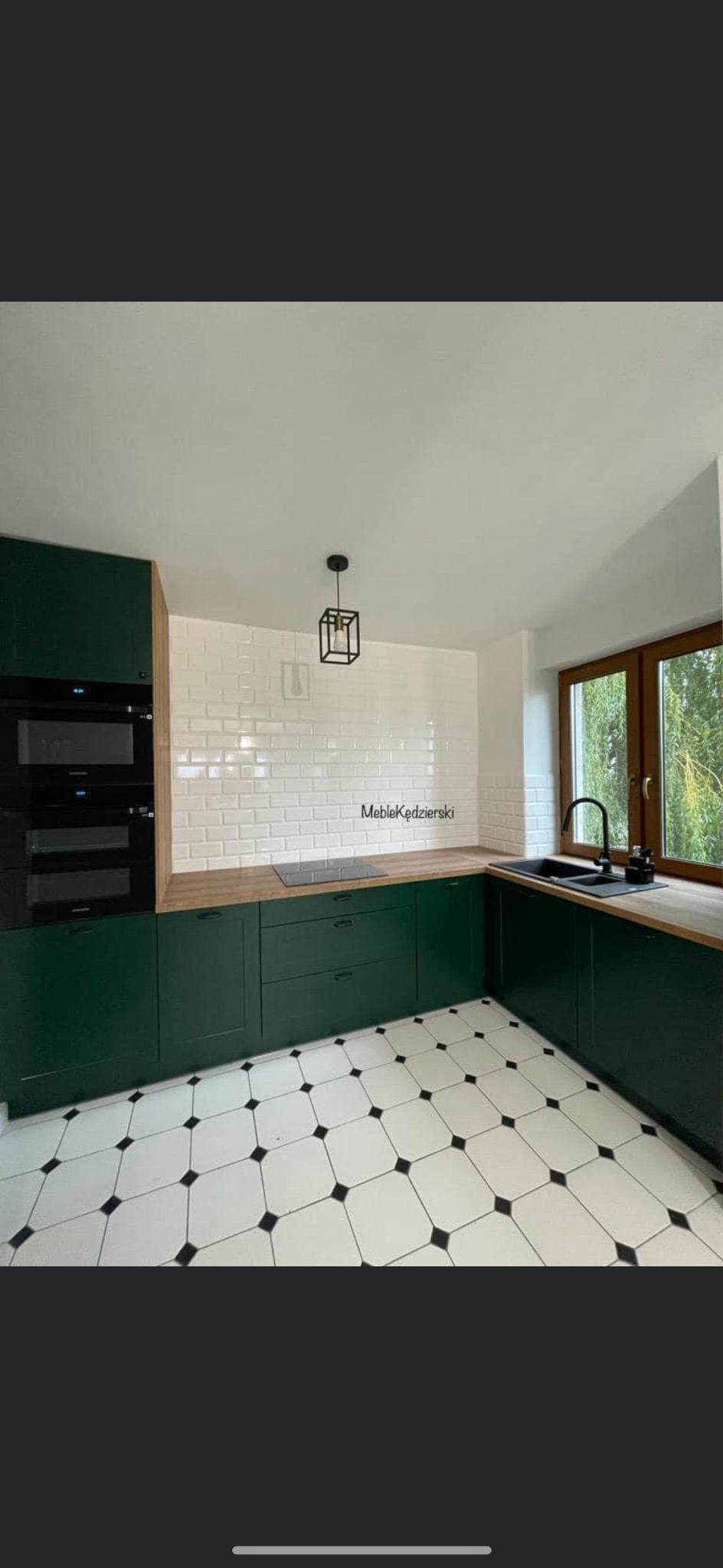 Nowoczesna kuchnia z ciemnozielonymi szafkami, drewnianym blatem i białą cegłą na ścianie, czarne dodatki i podłoga w biało-czarną szachownicę.