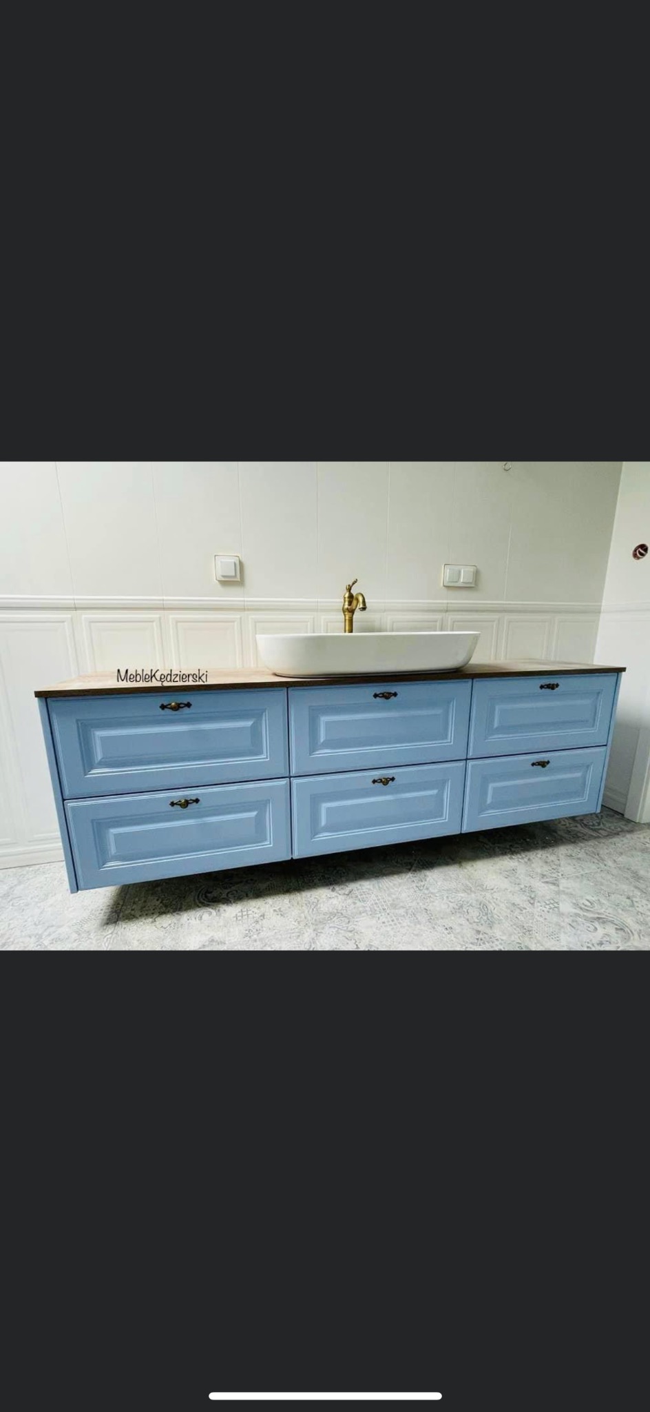 Niebieska szafka łazienkowa pod umywalkę nablatową z drewnianym blatem i mosiężną baterią, wisząca na ścianie z białą boazerią.