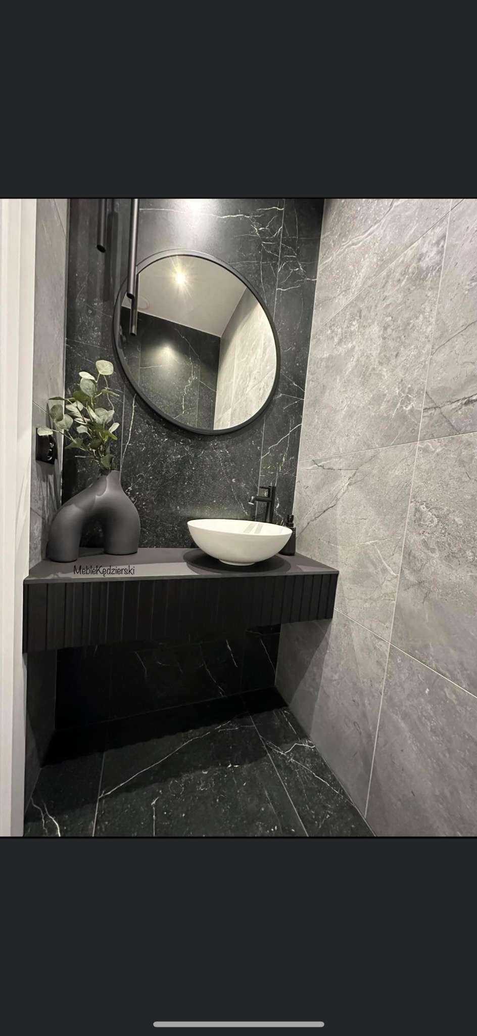 Nowoczesna łazienka z czarnymi marmurowymi ścianami i podłogą, białą umywalką nablatową, czarną armaturą i wiszącą szafką z ryflowanym frontem, uzupełniona okrągłym lustrem i dekoracyjnym wazonem...