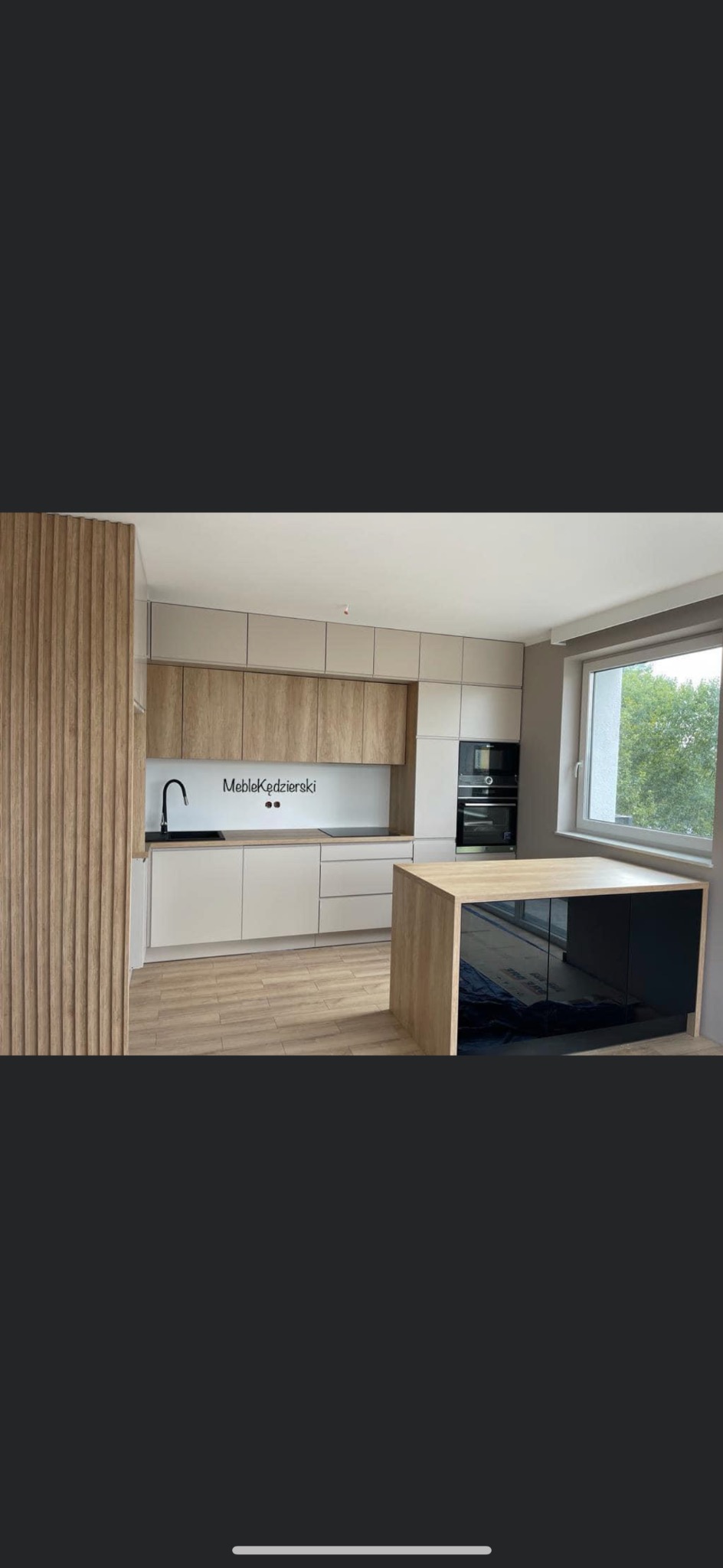 Nowoczesna kuchnia z wyspą, fronty w kolorze beżowym i drewnopodobnym, czarny zlew i płyta indukcyjna, drewniana lamela na ścianie, duże okno z widokiem na zieleń.