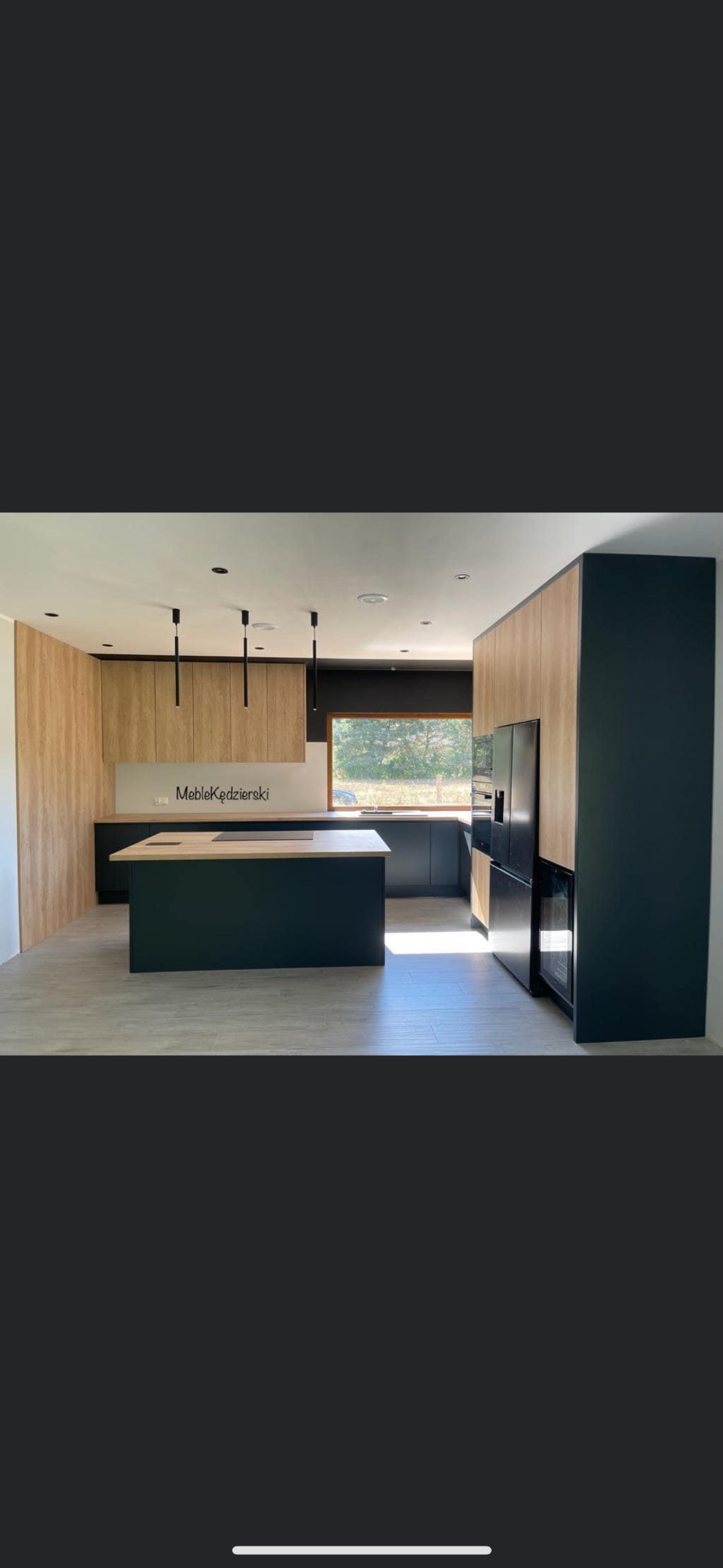 Nowoczesna kuchnia z wyspą, drewnianymi frontami szafek i czarnymi elementami, oświetlona naturalnym światłem z okna, z widocznym logo 'MebleKędzierski' na tle szafek.