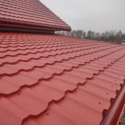 Czerwony dach z blachodachówki modułowej, widok z góry, z zamontowanymi rynnami w kolorze dachu i elementami mocującymi. W tle drzewa i pochmurne niebo.