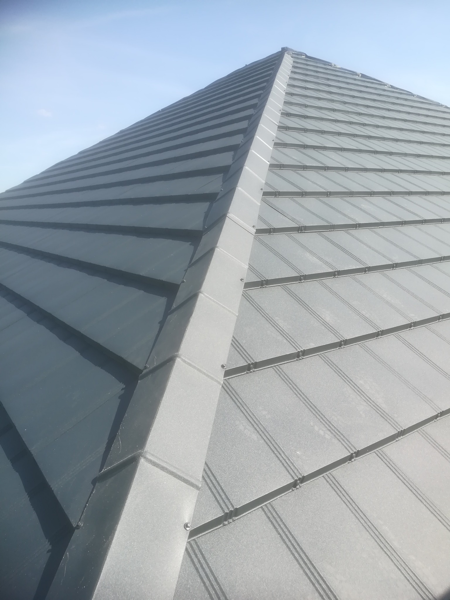 Metalowy dach w kolorze grafitowym, widok z dołu na szczyt dachu, z widocznymi panelami i obróbkami blacharskimi, na tle jasnego nieba.