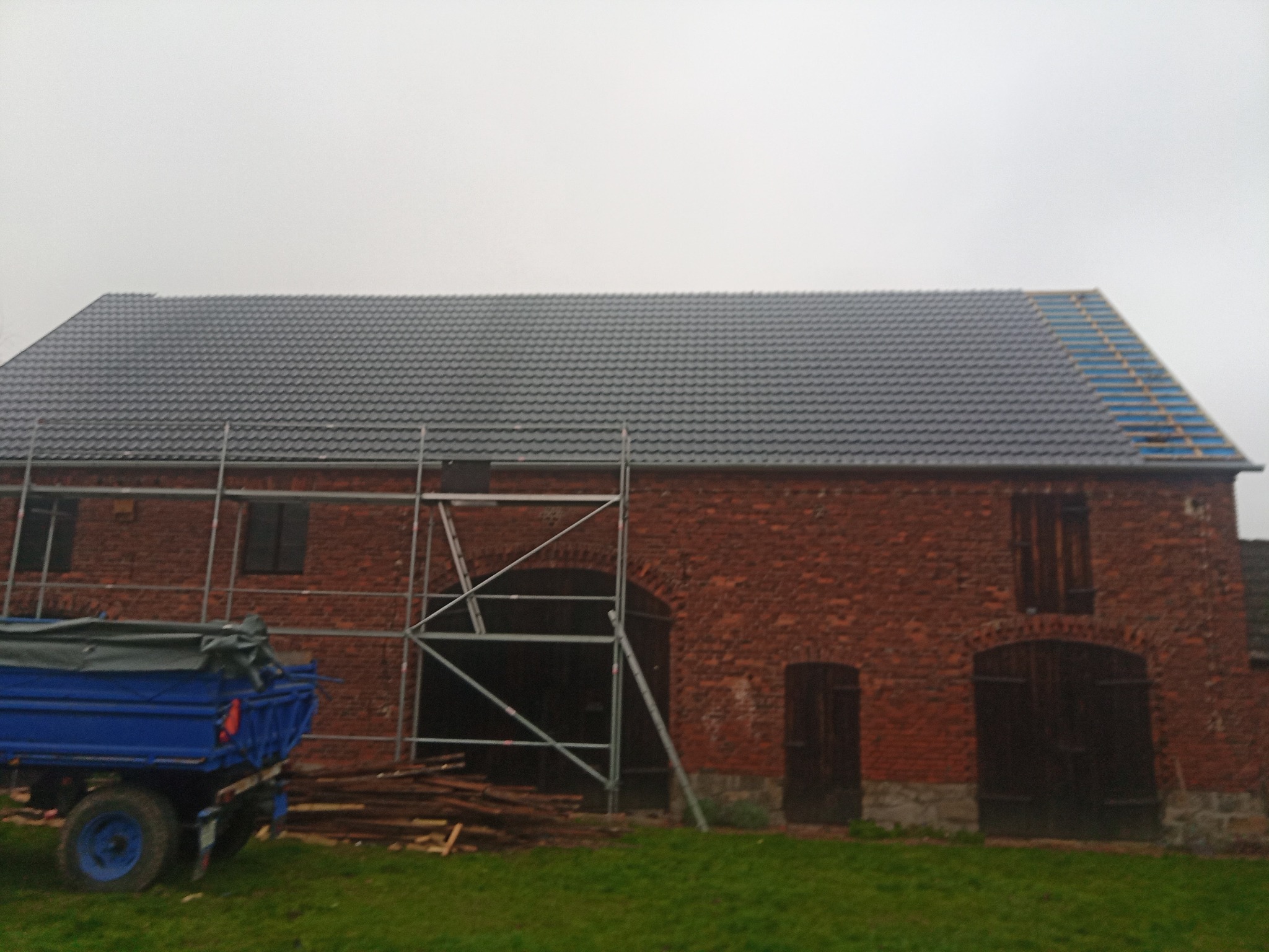 Częściowo wymieniony dach z szarej dachówki na ceglanym budynku gospodarczym, widoczne rusztowanie i przyczepa rolnicza z plandeką.