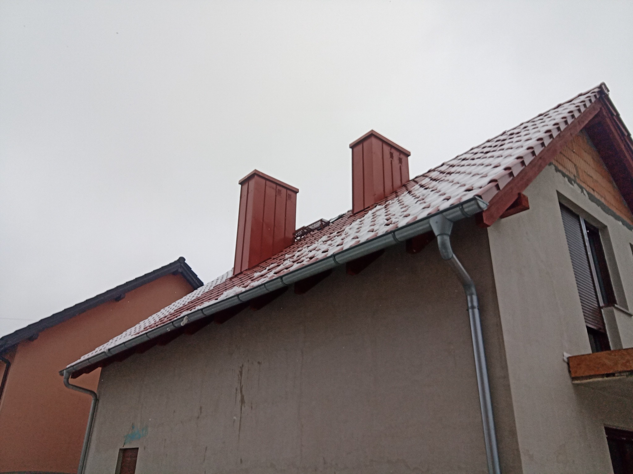 Dom z czerwonym dachem pokrytym lekkim śniegiem, widoczne dwa kominy w kolorze dachu oraz rynny, pochmurne niebo w tle.