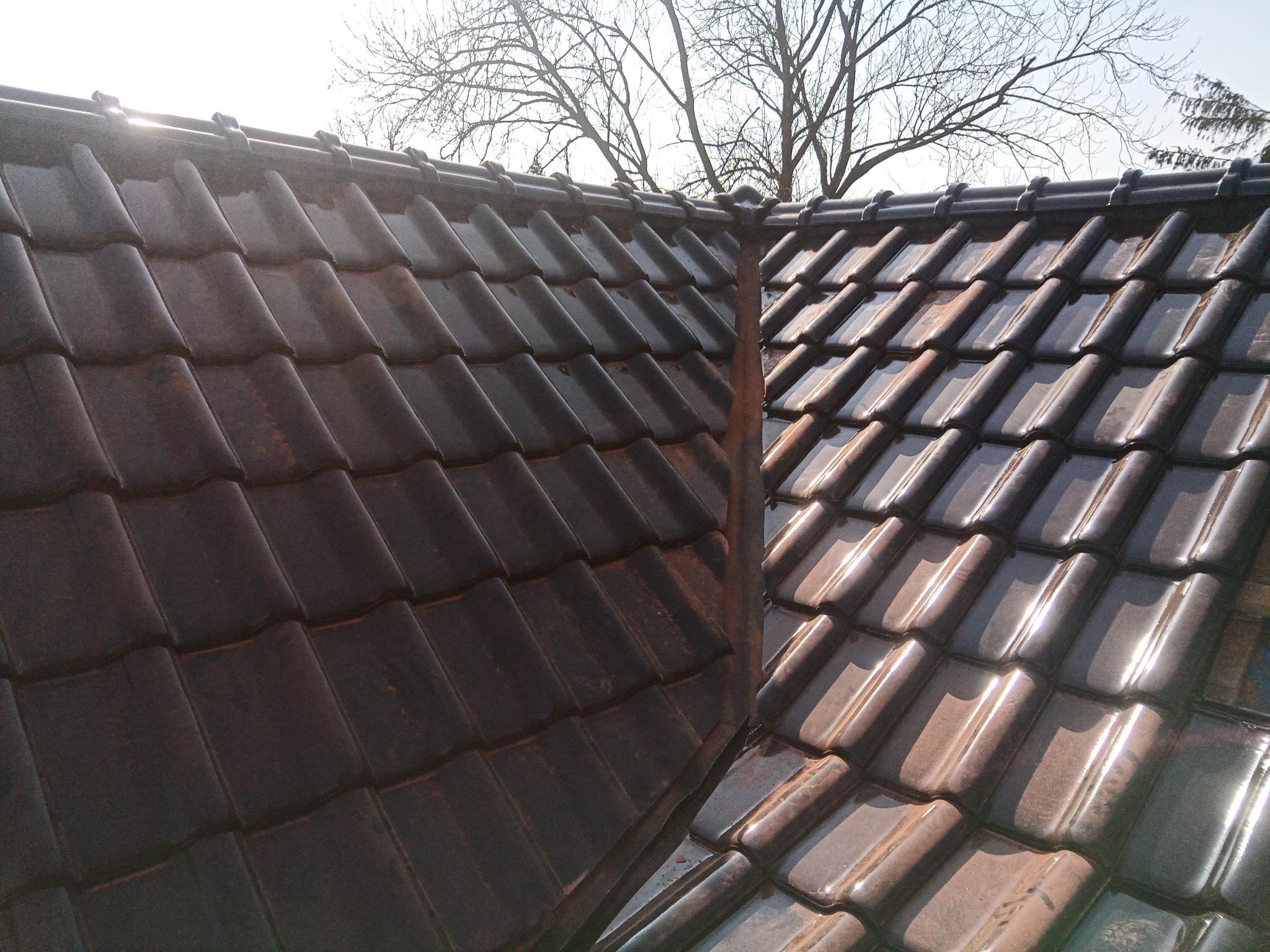Fragment dachu pokrytego ciemną dachówką ceramiczną, widoczne obróbki blacharskie w narożniku dachu, w tle drzewo bez liści i jasne niebo.