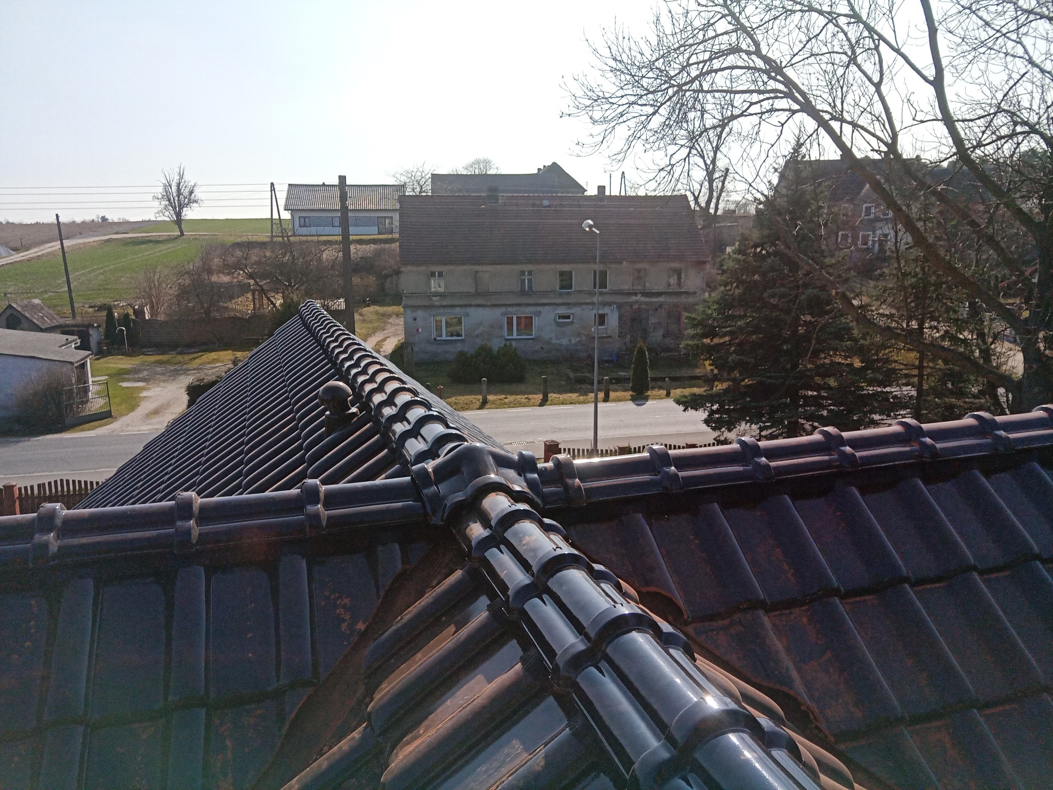 Nowy dach z błyszczącej, ciemnej dachówki ceramicznej w perspektywie z góry, z widokiem na wiejski krajobraz i zabudowania w tle.