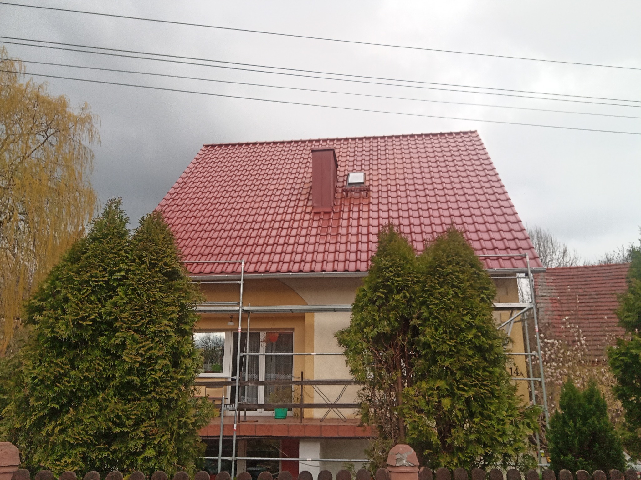Dom jednorodzinny z nowym, czerwonym dachem z dachówki ceramicznej, częściowo zasłonięty przez rusztowanie i wysokie tuje, widoczne przewody elektryczne na tle pochmurnego nieba.