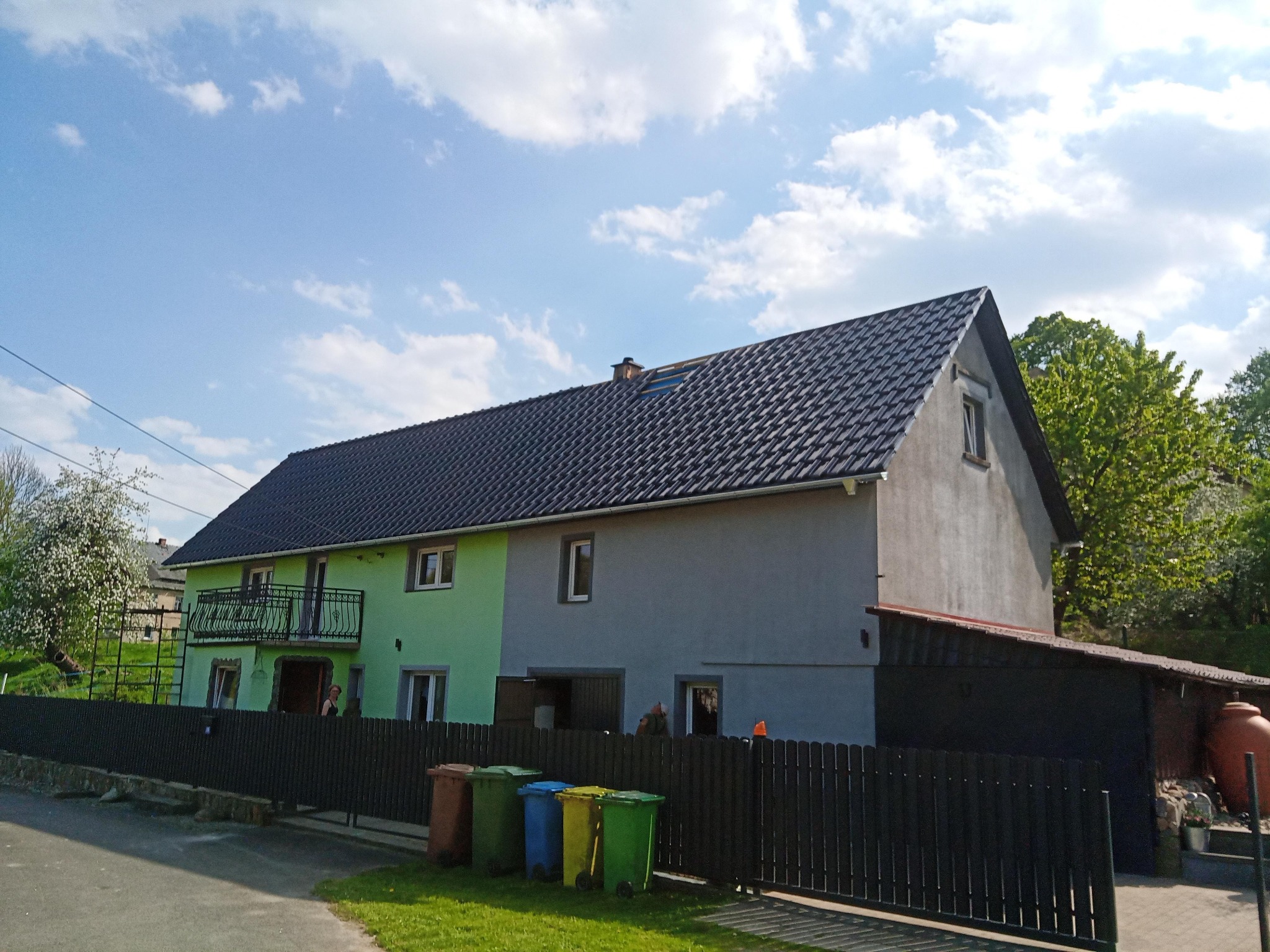 Dwukondygnacyjny budynek z nowym, ciemnoszarym dachem z dachówki ceramicznej, widoczny komin i okno dachowe, elewacja w dwóch odcieniach zieleni i szarości, czarne ogrodzenie, kolorowe pojemniki...
